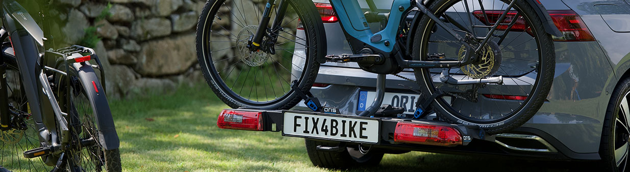 ORIS TRACC FIX4BIKE bicycle carrier mounted on a grey Volkswagen Golf 8 with two e-bikes, shown on a grassy area with stone wall and parked bicycle in the foreground.