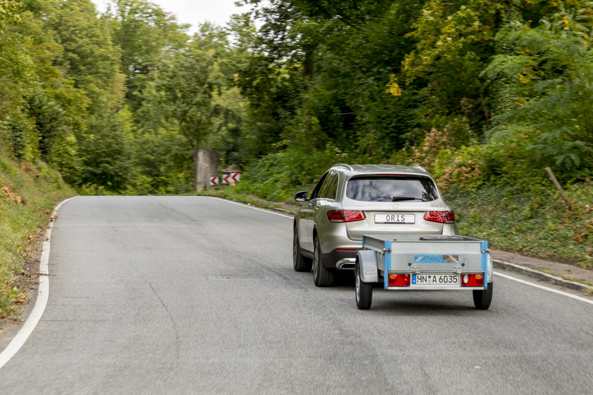 Car towing a small utility trailer with an ORIS trailer hitch while driving on a winding country road surrounded by dense green trees.