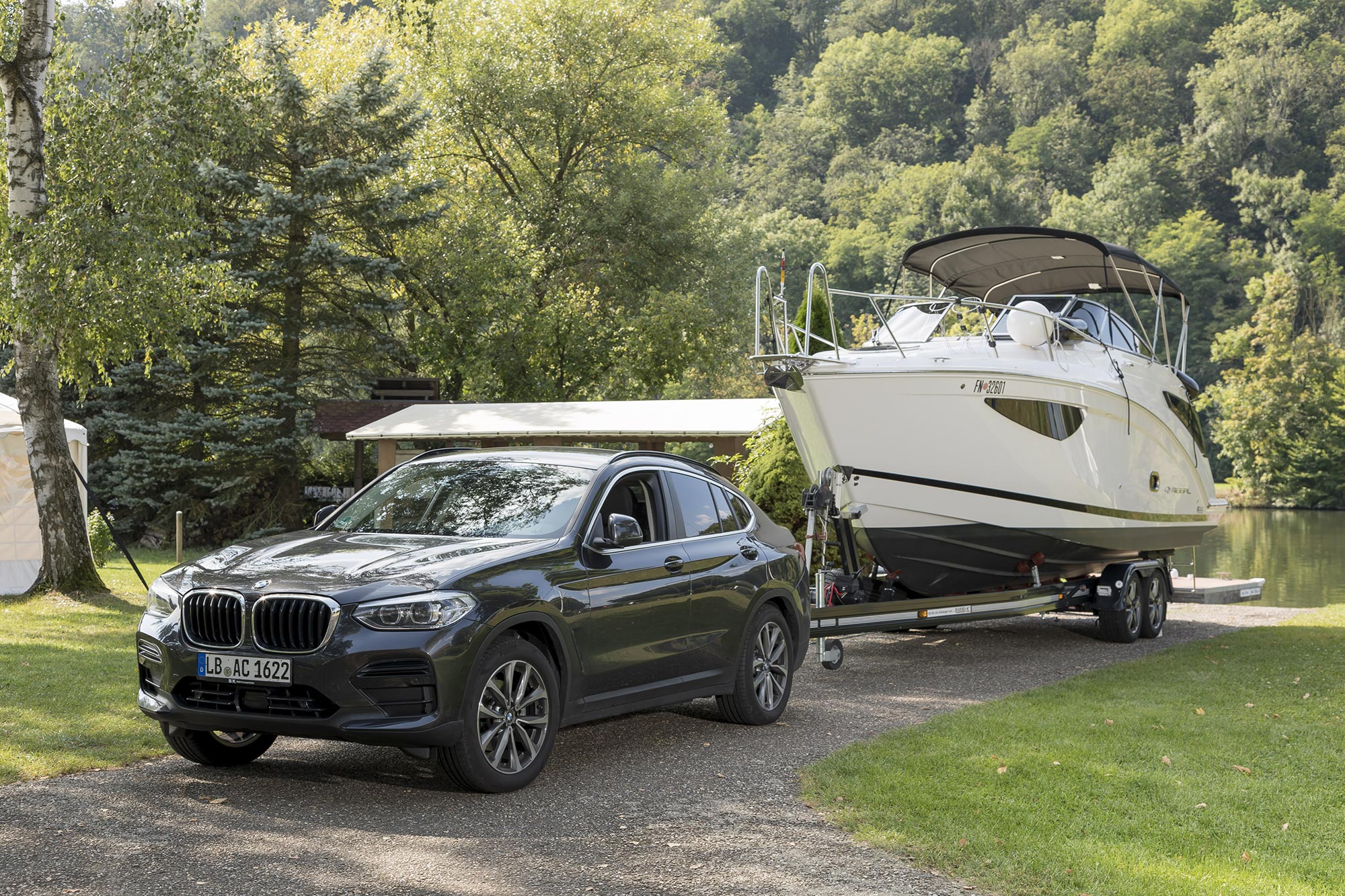 Black BMW X4 towing a motorboat on a trailer near a lakeside campsite surrounded by trees and green landscape.