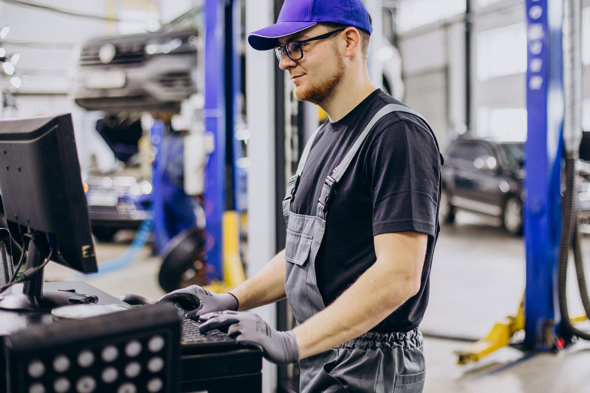 Service technician operating a computer workstation in an automotive workshop with vehicles on lifting platforms in the background.