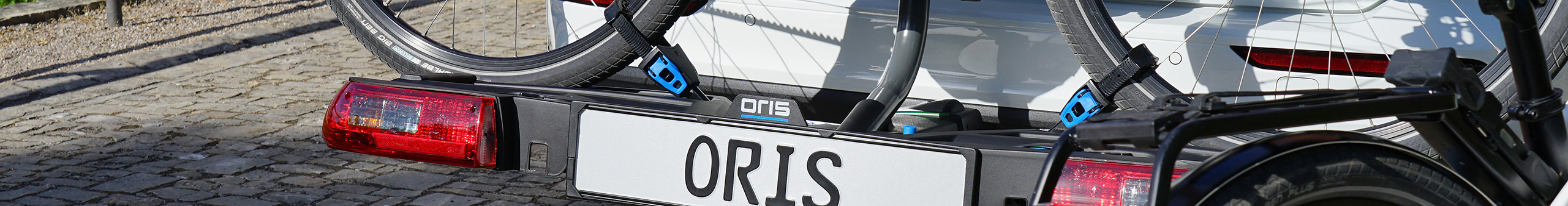 Close-up of ORIS TRACC bicycle carrier mounted to a white Volvo estate car, showing integrated rear lights, number plate holder and wheel straps securing an e-bike, with a second e-bike visible in the foreground on cobblestone pavement.