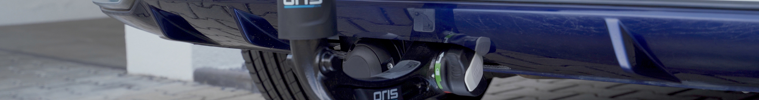 Detailaufnahme einer ORIS AK 4 FIX4BIKE® vertikal abnehmbaren Anhängerkupplung mit Schutzkappe und Steckdose an einem blauen Fahrzeug montiert.