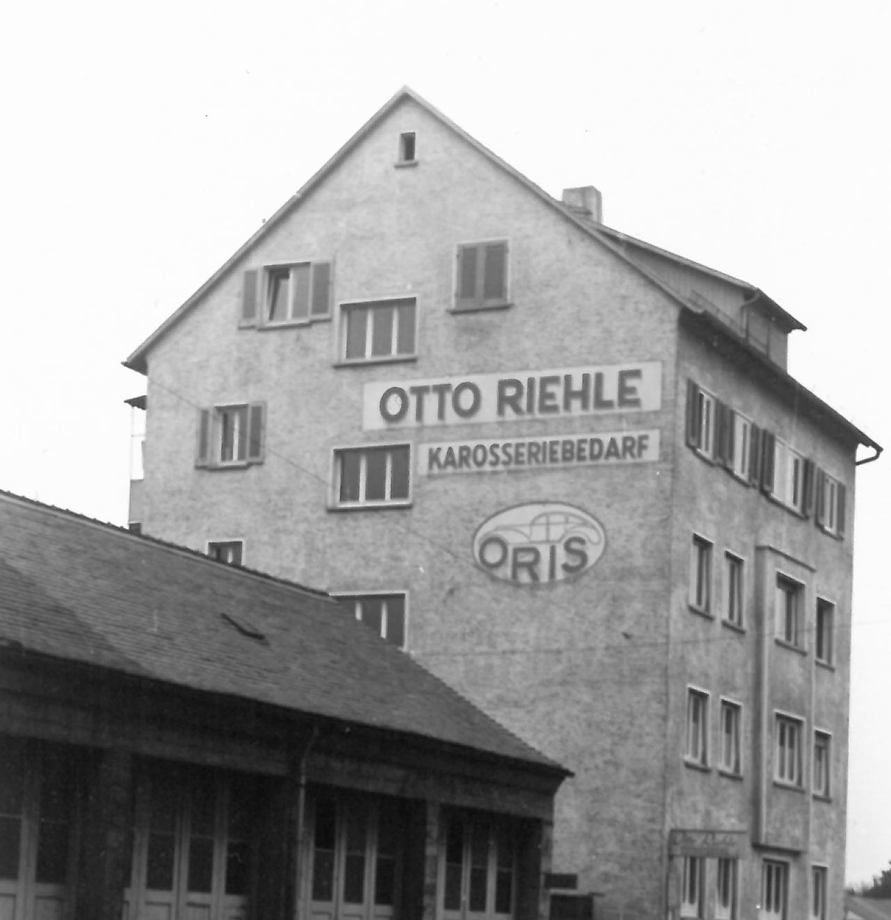 Historic black-and-white photograph of the original ORIS company building in Stuttgart, featuring the Otto Riehle Karosseriebedarf signage and ORIS logo on the facade.