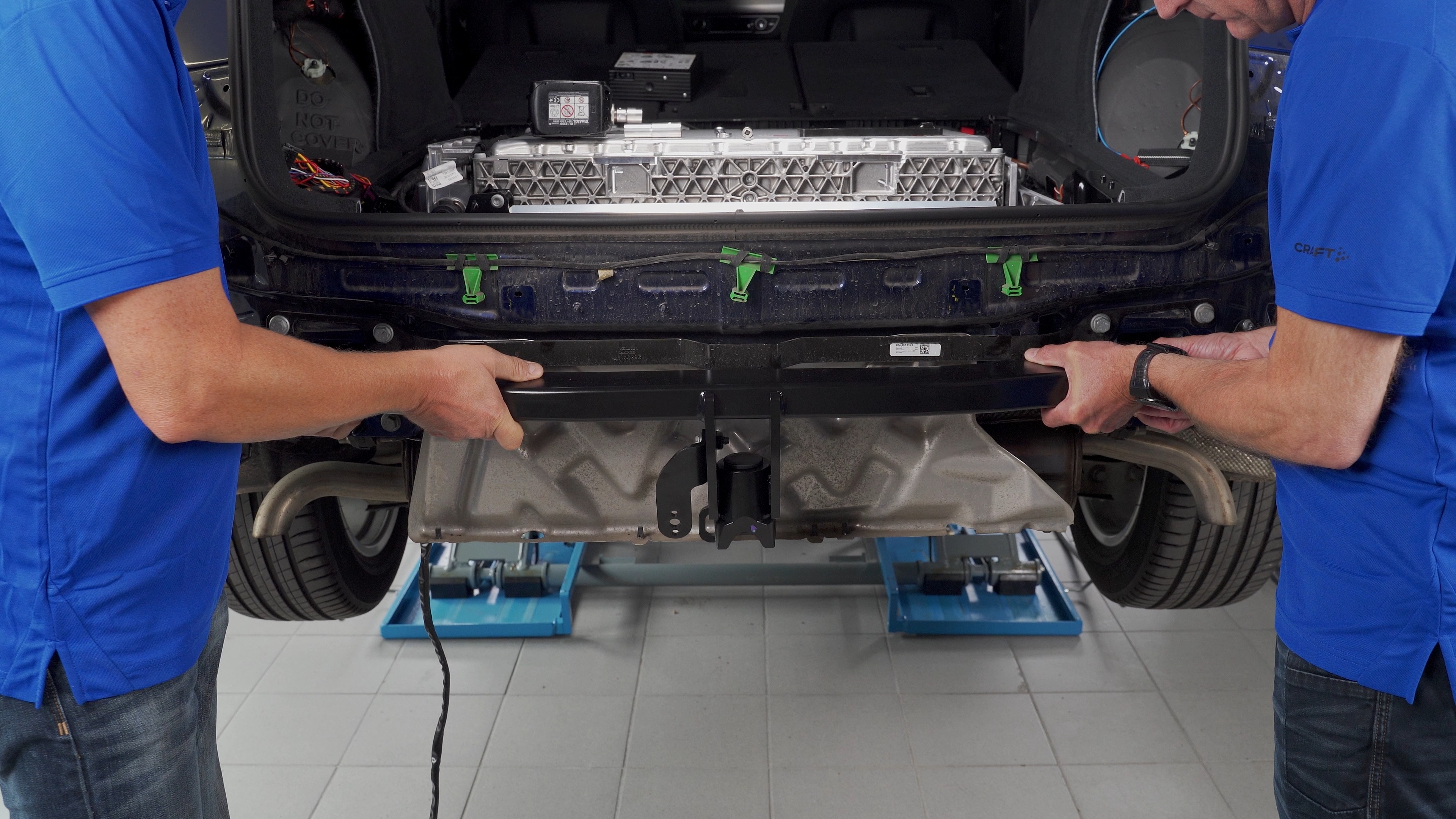 Two technicians positioning an ORIS towbar crossmember on a blue Audi during towbar installation, with the high-voltage battery visible in the luggage compartment.