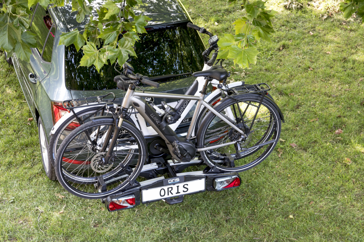 ORIS TRACC bicycle carrier mounted on a towbar carrying two e-bikes on the rear of a car parked on grass beneath green trees.