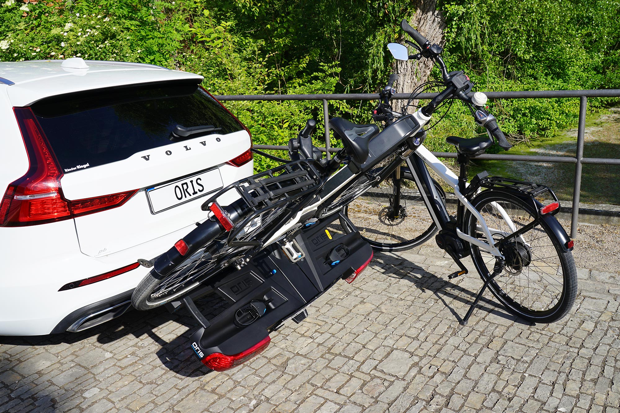 ORIS TRACC bicycle carrier mounted to a white Volvo estate car with tilted rear access function, carrying one e-bike while a second e-bike stands on the ground beside the vehicle, surrounded by trees, grass and a green park landscape.