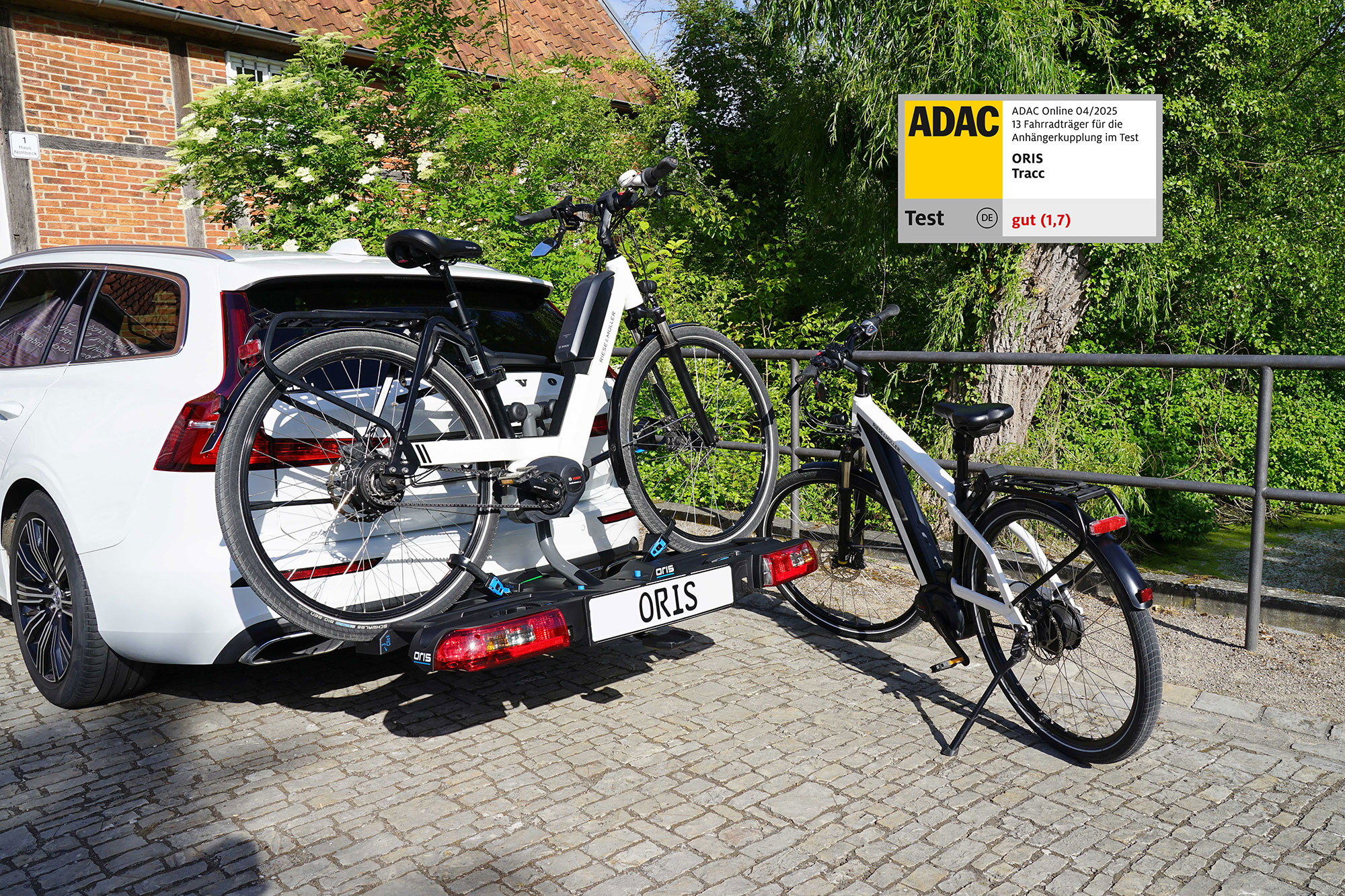 ORIS Tracc FIX4BIKE® bike carrier mounted on a vehicle towbar with one e-bike loaded and a second e-bike positioned beside the vehicle, featuring an ADAC test rating graphic in the upper right corner.