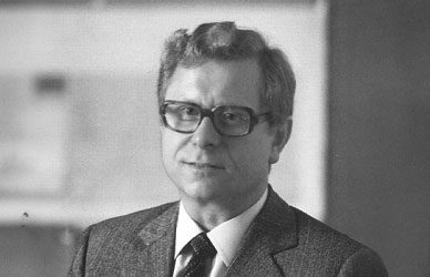 Black-and-white portrait of Hans Riehle wearing a suit, tie and glasses, looking toward the camera against a blurred indoor background.
