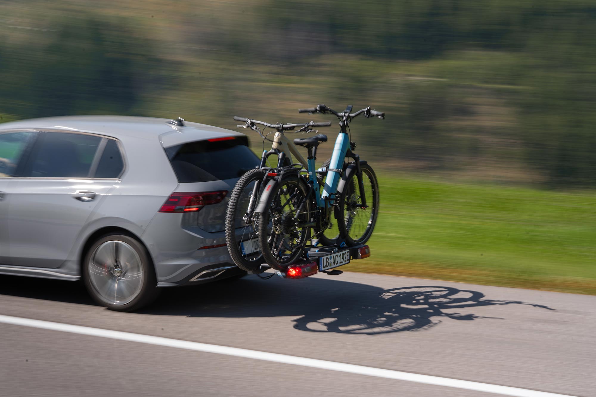 Grey Volkswagen Golf 8 driving with TRACC bicycle carrier carrying two e-bikes on a country road with motion blur and green landscape in the background.