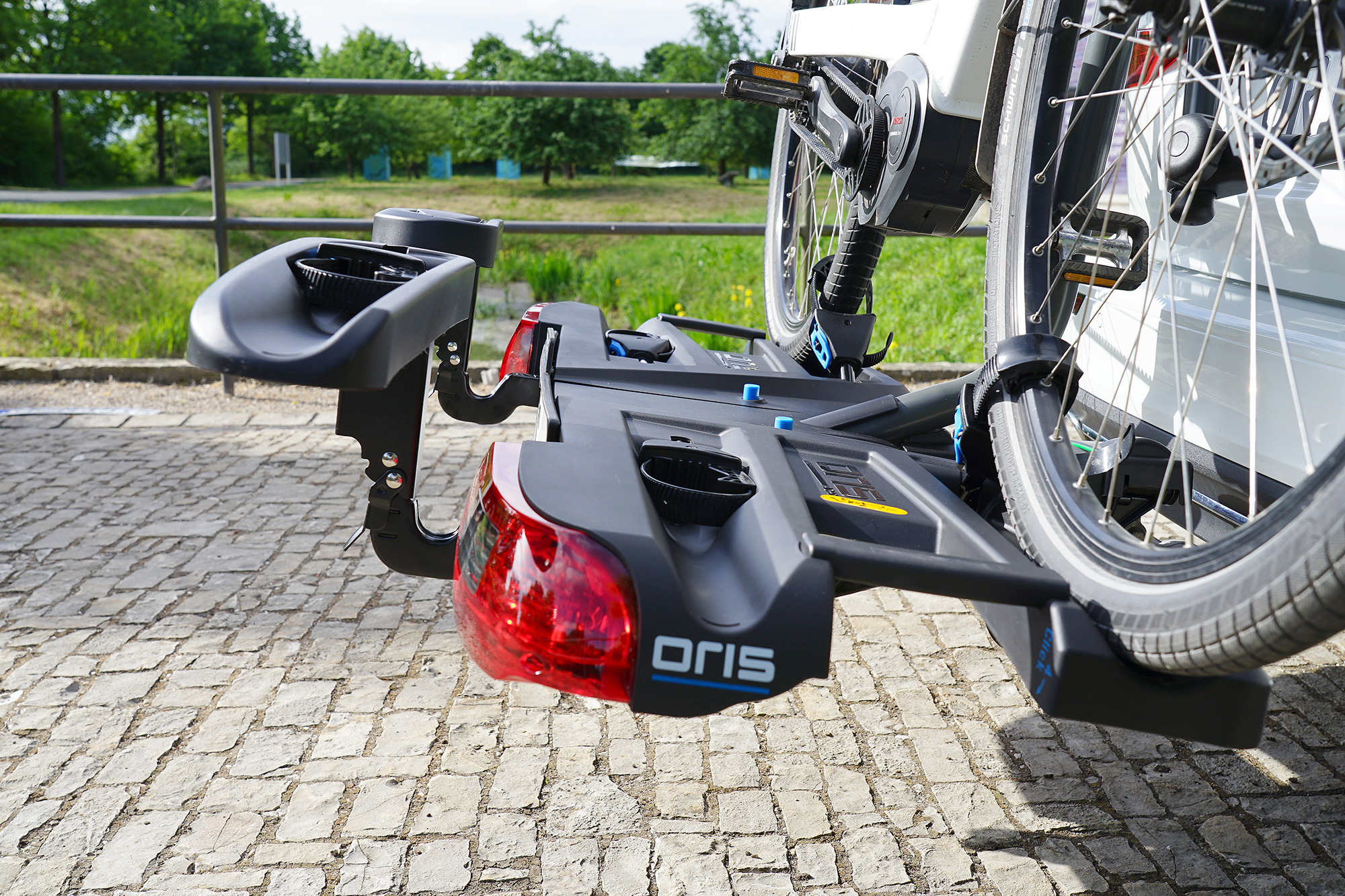 Close-up of the ORIS TRACC FIX4BIKE bicycle carrier with mounted e-bike and installed extension rail for carrying an additional bicycle, featuring wide wheel trays on a white vehicle beside a green riverside landscape.
