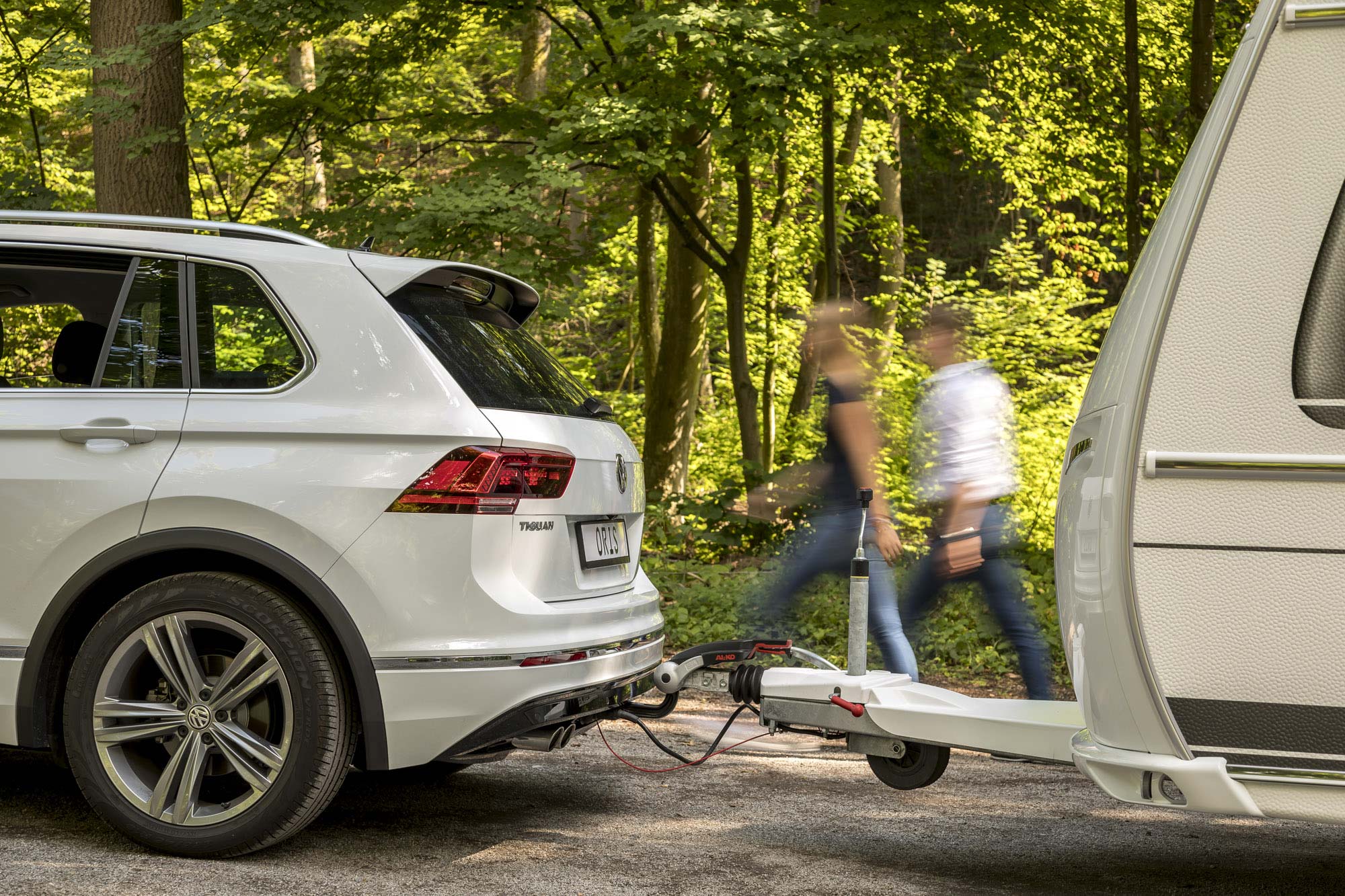 White Volkswagen Tiguan with towbar coupled to a caravan on a forest road, with two blurred people walking in the background.