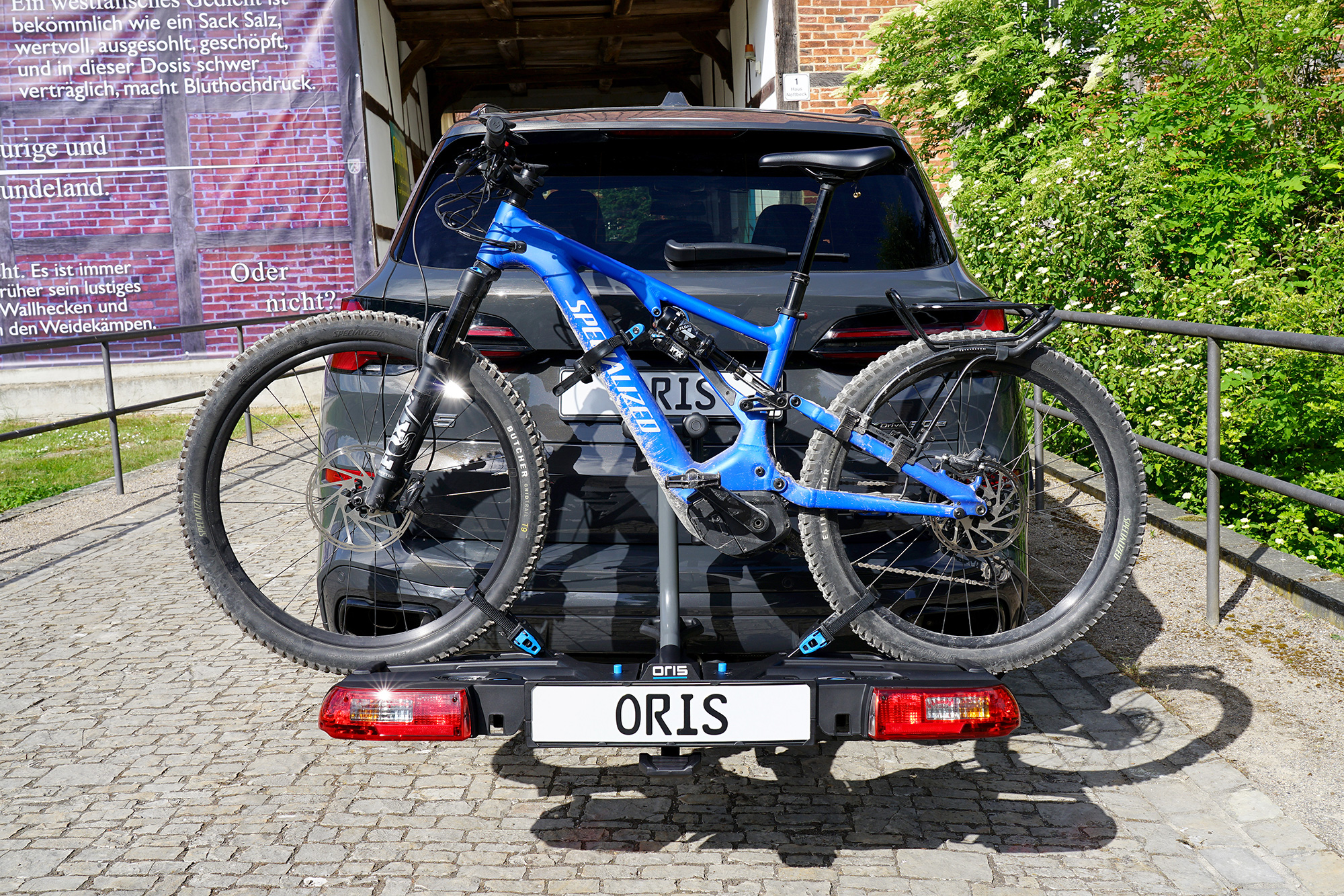 Blue Specialized mountain bike mounted on the ORIS TRACC FIX4BIKE bicycle carrier attached to a dark grey BMW X5, parked on a cobblestone area between historic brick buildings, greenery and a riverside railing.