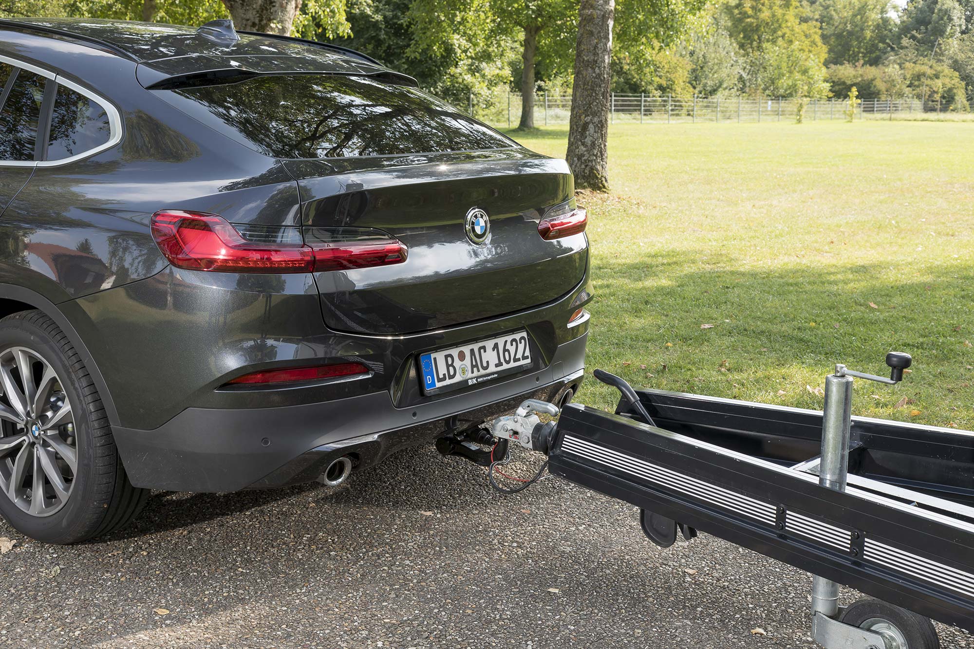 Rear view of a dark BMW X4 with a trailer coupled to the towbar on a gravel area beside trees and a meadow.