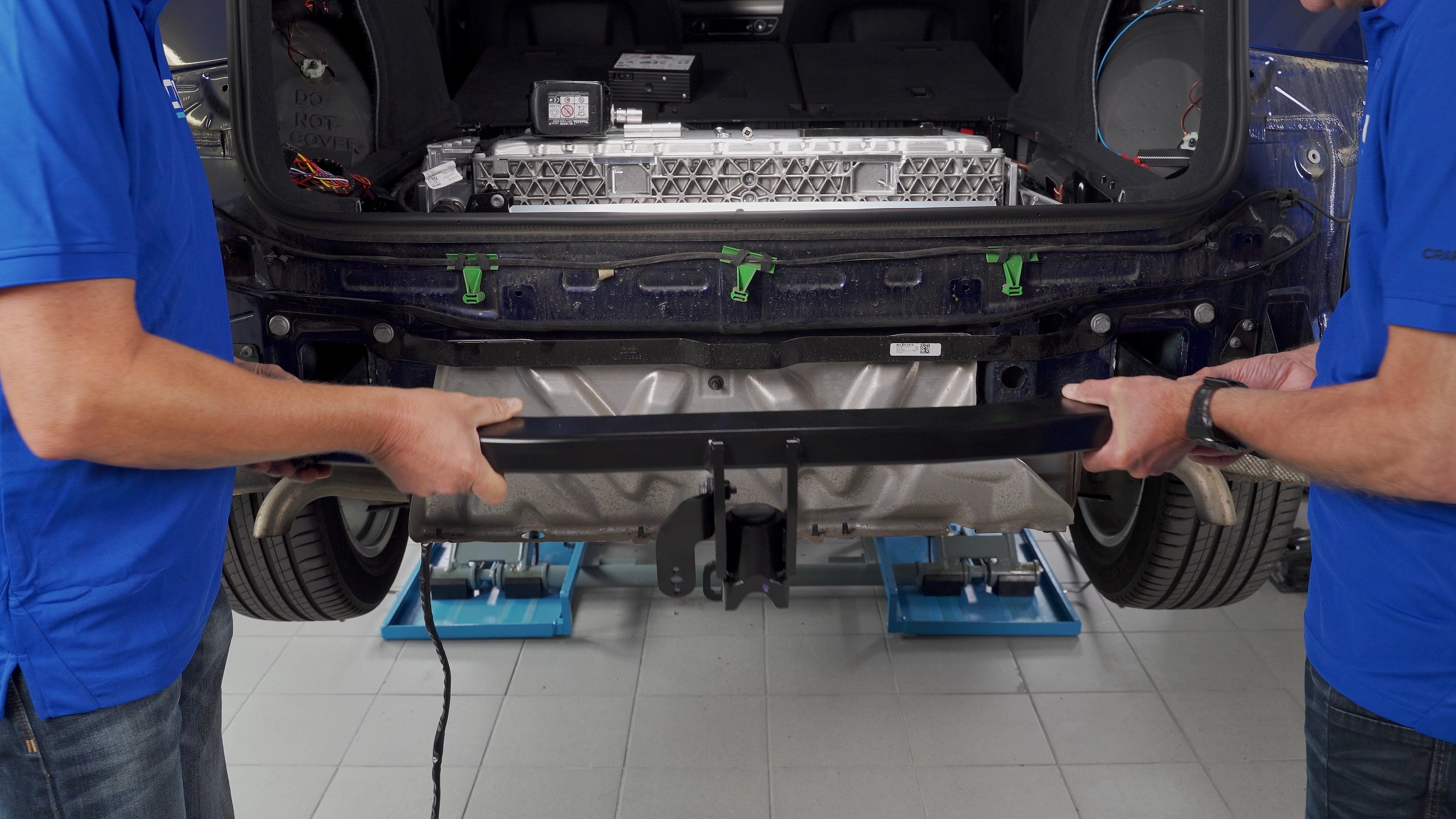 Two technicians positioning an ORIS towbar crossmember on a blue Audi during towbar installation, with the high-voltage battery visible in the luggage compartment.