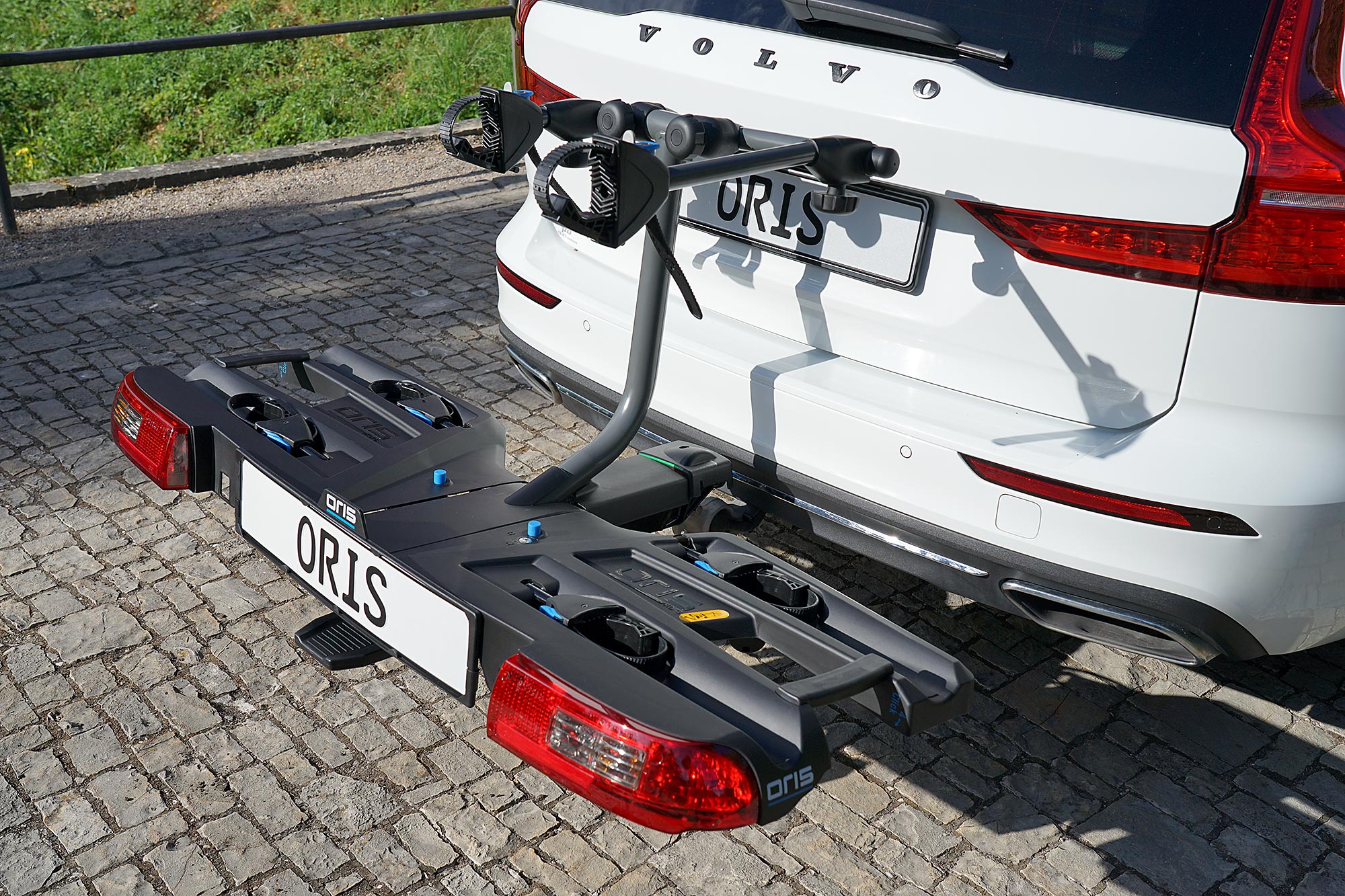 ORIS TRACC bicycle carrier with high load capacity mounted on a white Volvo, featuring four wheel trays and adjustable frame holders on a cobblestone riverside promenade.