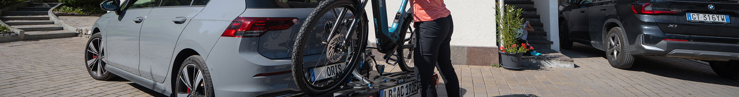 Person lädt ein E-Bike auf einen ORIS FIX4BIKE Fahrradträger an einem grauen Volkswagen Golf 8, geparkt auf einer Hofeinfahrt mit BMW SUV und Dachbox im Hintergrund.