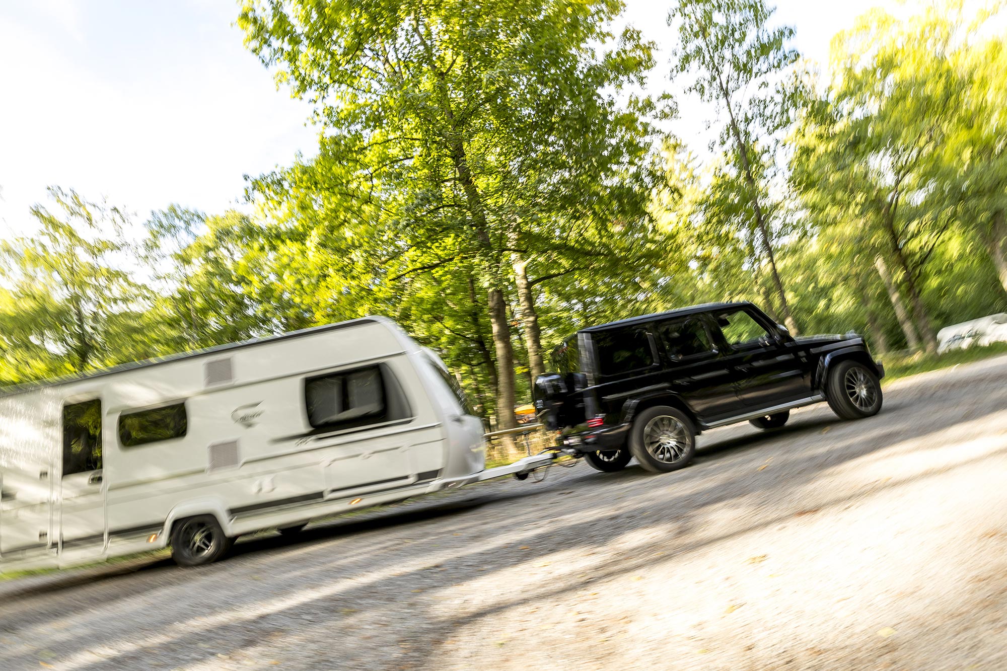 Black Mercedes-Benz G-Class towing a white caravan on a campsite road surrounded by trees and green woodland scenery.