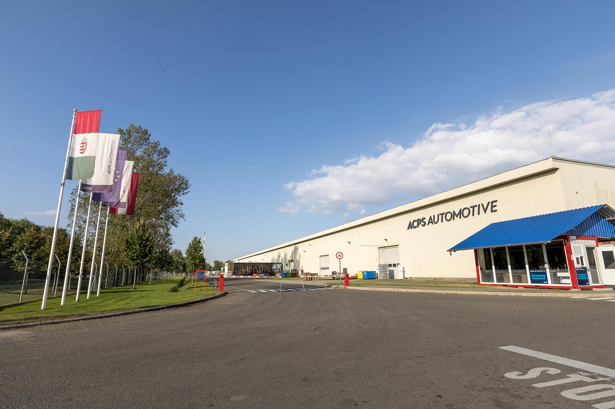 ACPS Automotive production facility in Kecskemét, Hungary, with manufacturing hall, company signage, entrance area and national flags along the access road.