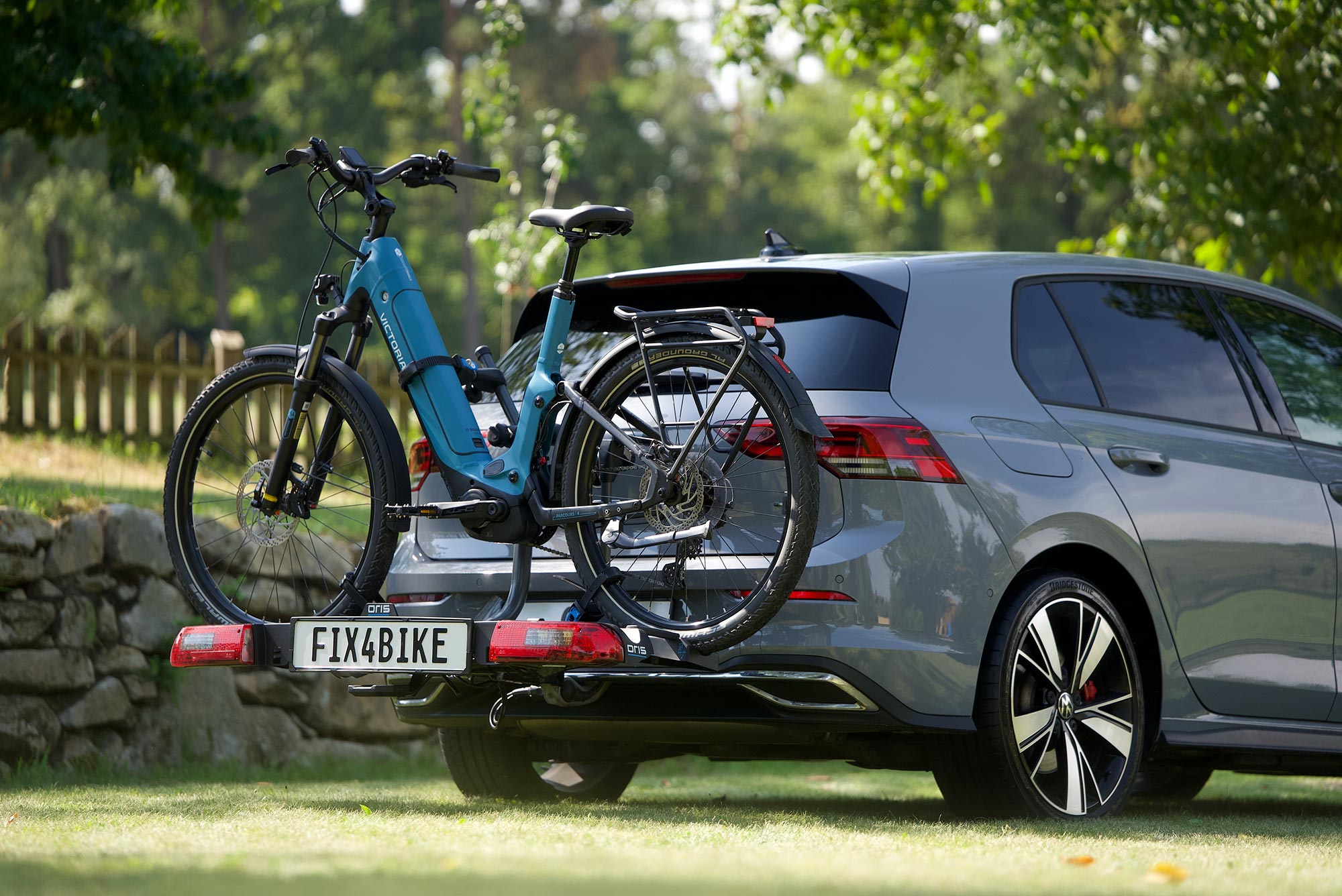 Grey Volkswagen Golf 8 with FIX4BIKE bicycle carrier transporting a blue e-bike on a grassy area surrounded by trees and park landscape.