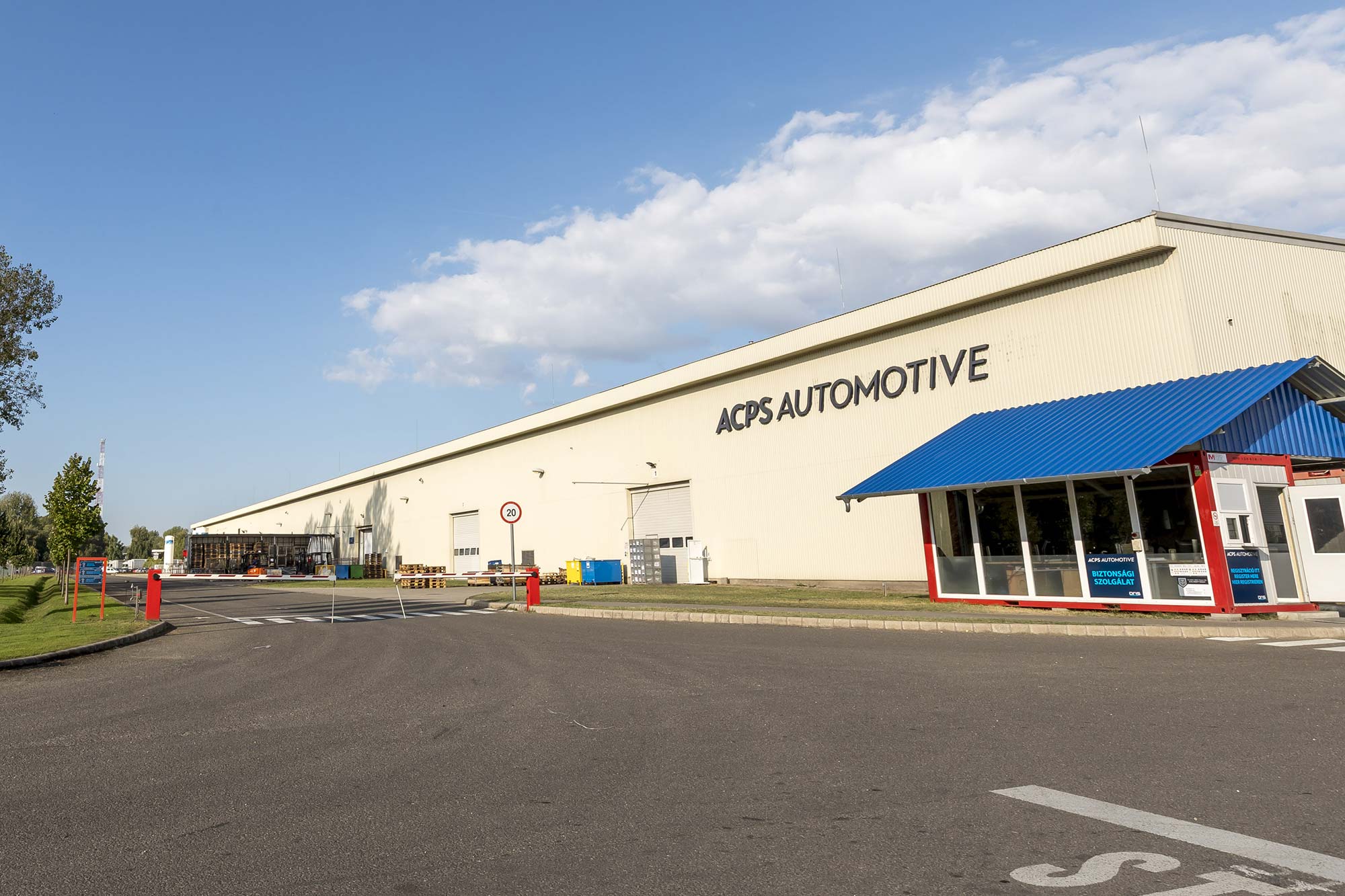ACPS Automotive building at the Hungary location with large warehouse facade, blue entrance canopy and open yard under a blue sky.