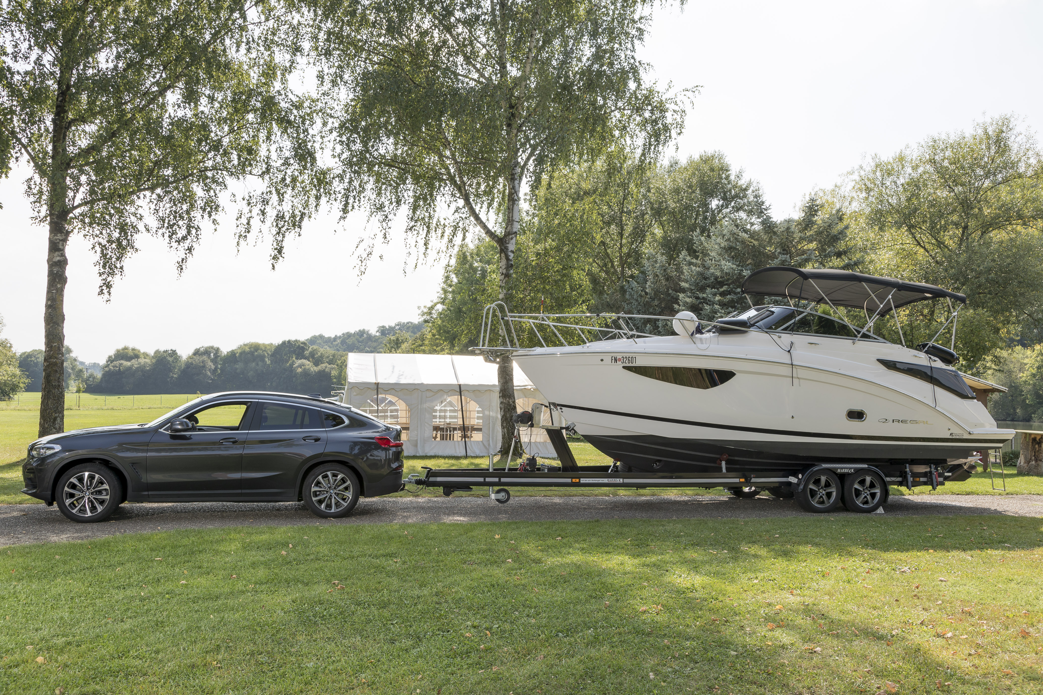 Side view of a black BMW SUV towing a large white motorboat on a tandem-axle trailer with an ORIS towbar. The image shows the complete vehicle and boat beside a grassy lakeside surrounded by trees.