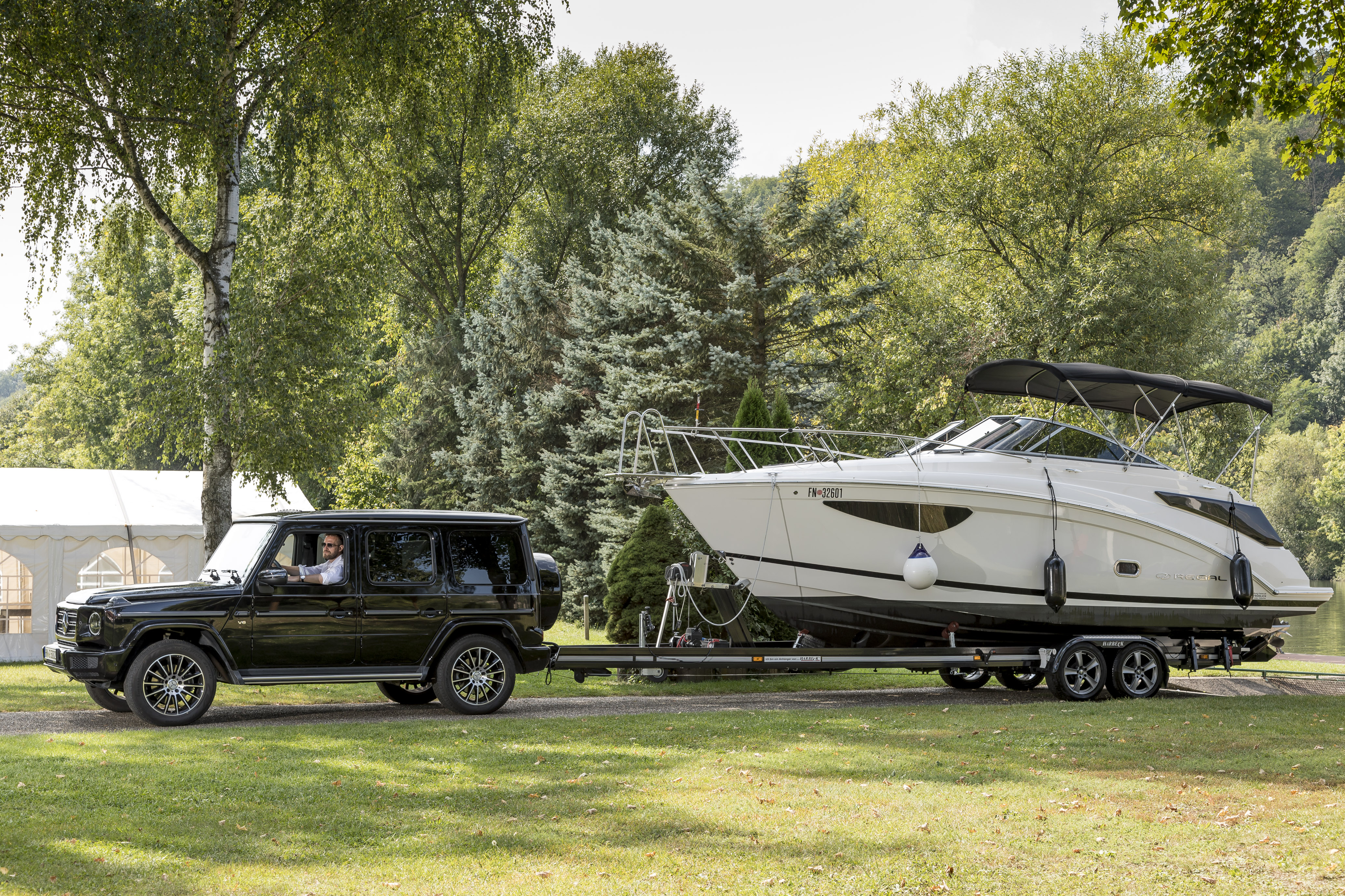 Black SUV towing a large motorboat on a trailer through a green park landscape with trees and lawn.