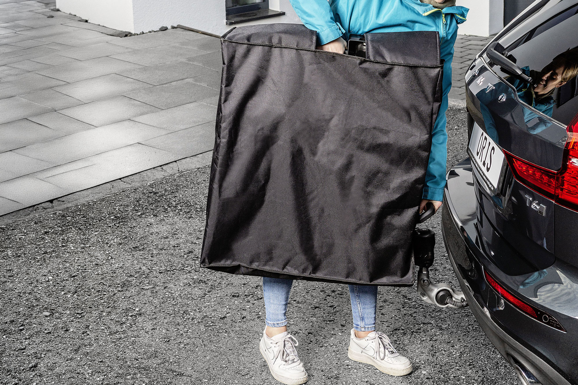 Person mit einer ORIS TRACC FIX4BIKE® Aufbewahrungstasche neben einem schwarzen Volvo mit montierter FIX4BIKE® Anhängerkupplung auf einer Einfahrt.