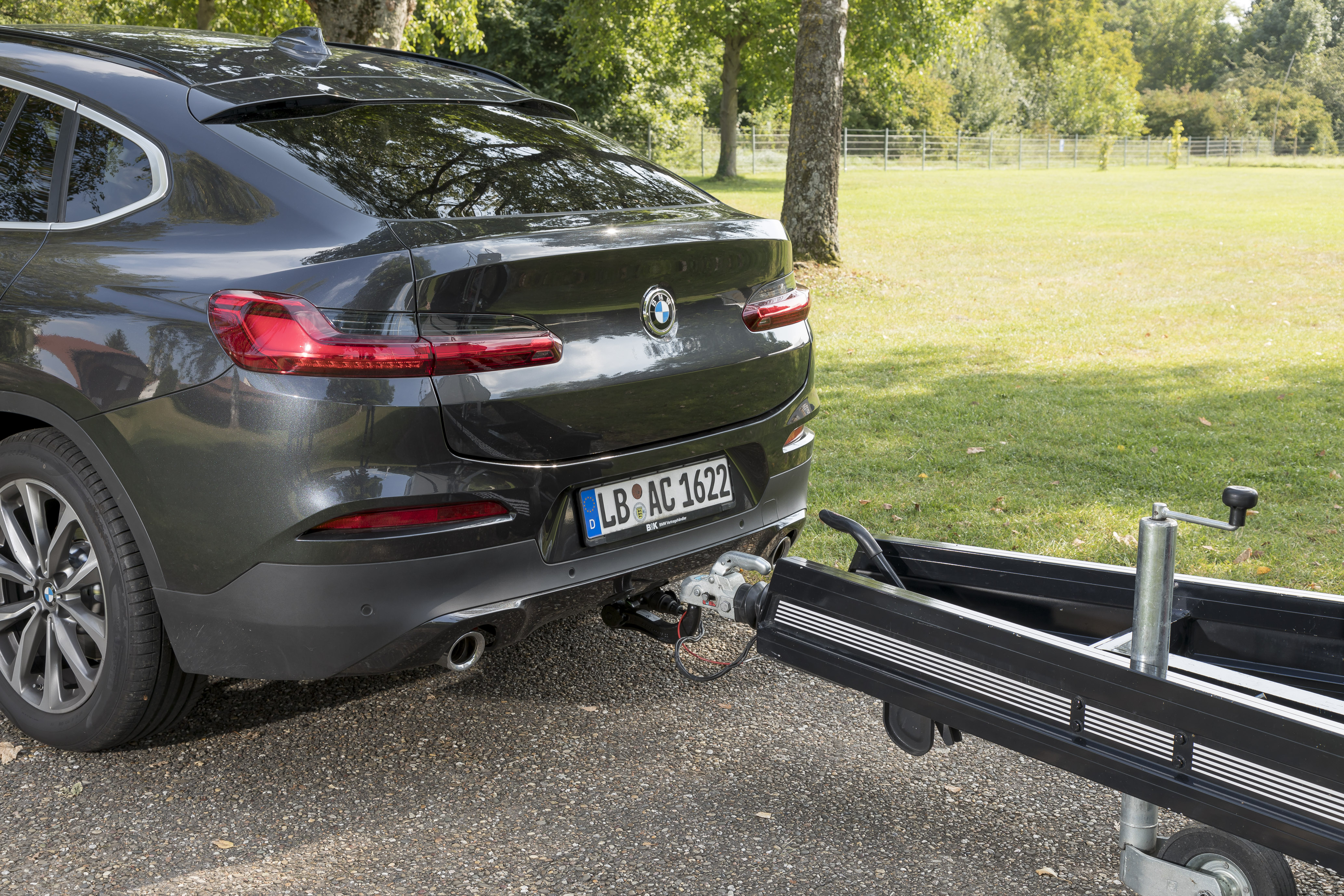Rear view of a BMW SUV with an ORIS towbar coupled to a boat trailer. The trailer is electrically connected via the towbar socket, with a grassy park and trees in the background.