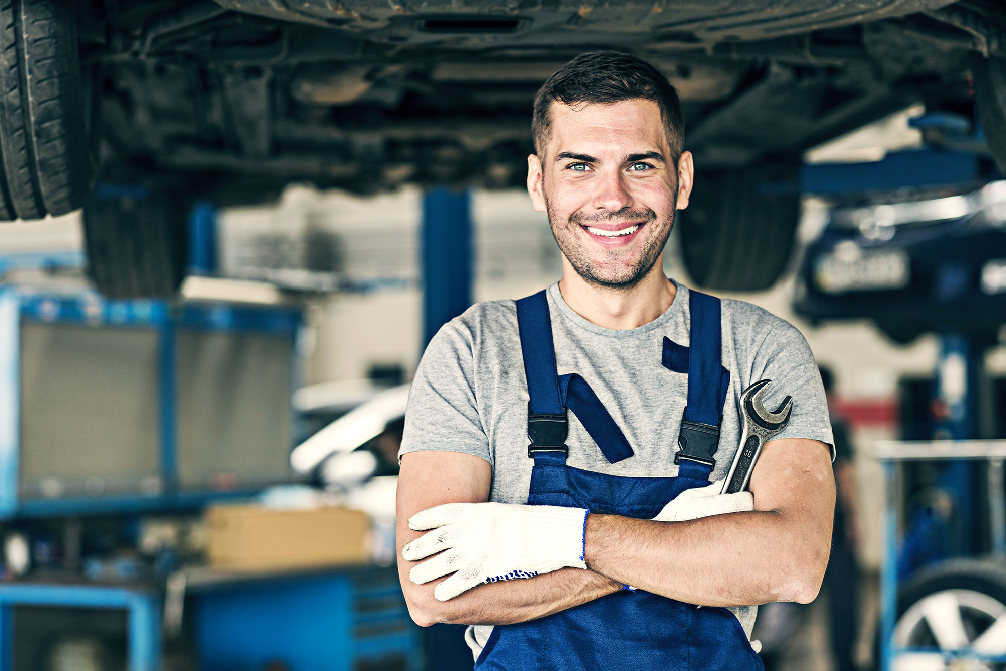 Smiling mechanic standing in a vehicle workshop holding a wrench in front of a raised car.