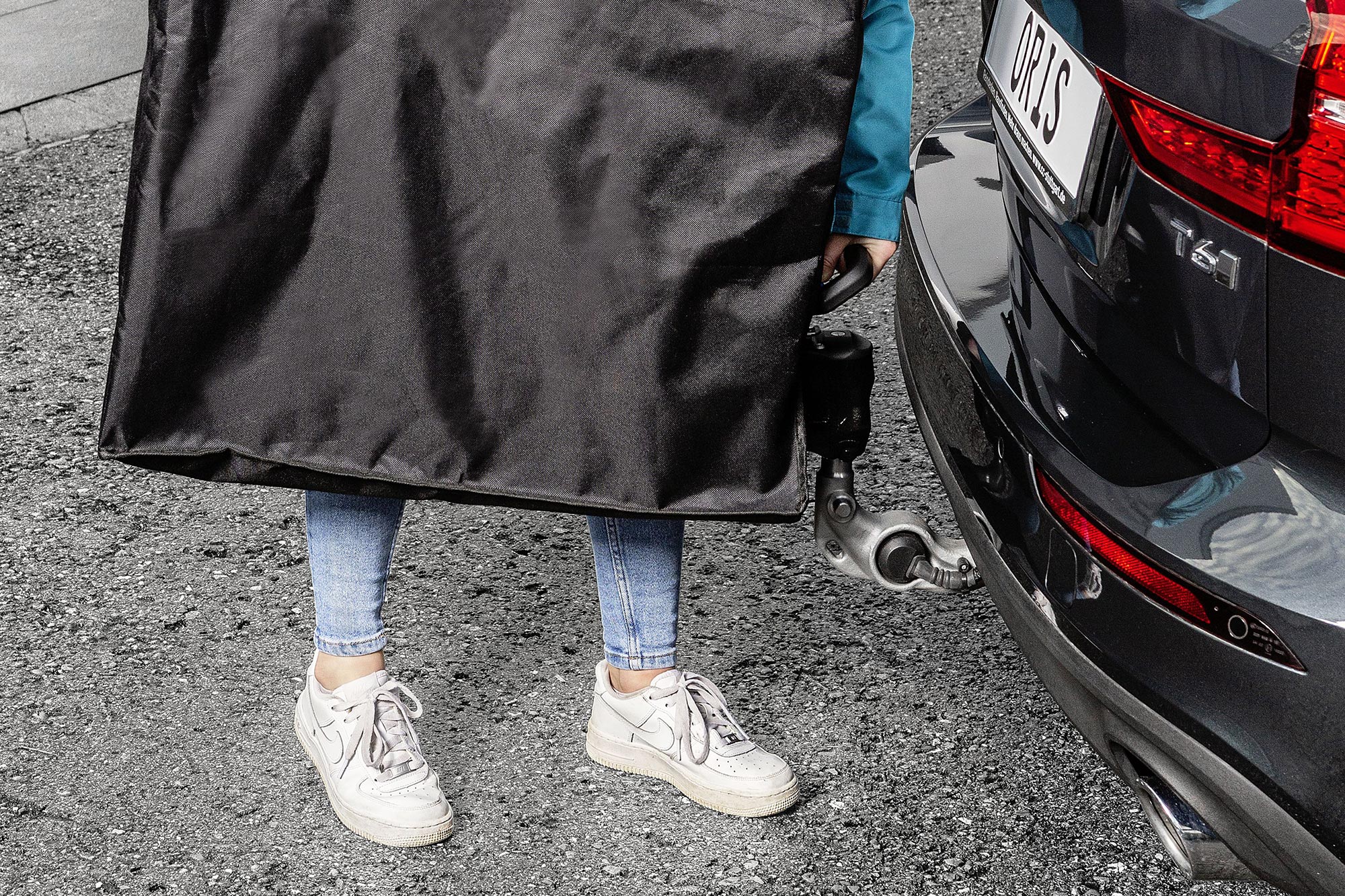Person carrying the folded ORIS TRACC FIX4BIKE bicycle carrier in a transport bag beside a black Volvo with electrically swivelling ORIS towbar.