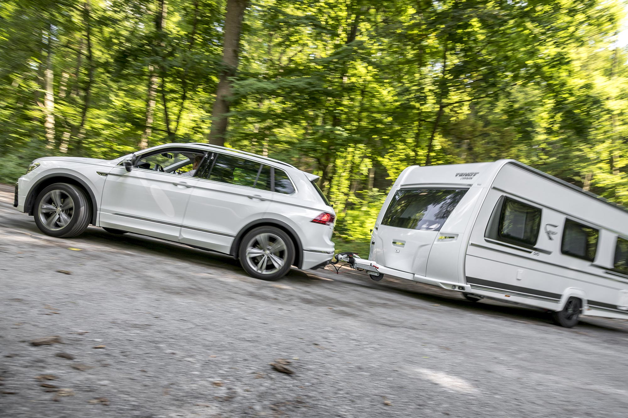 White Volkswagen Tiguan towing a white caravan on a forest road surrounded by trees and green woodland scenery.
