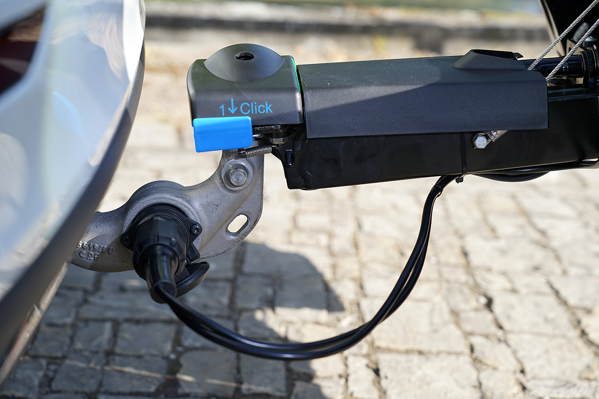Close-up of the ORIS TRACC Maximum Safety coupling connection with 1-Click locking mechanism and electrical plug mounted on a towbar above a cobblestone surface.