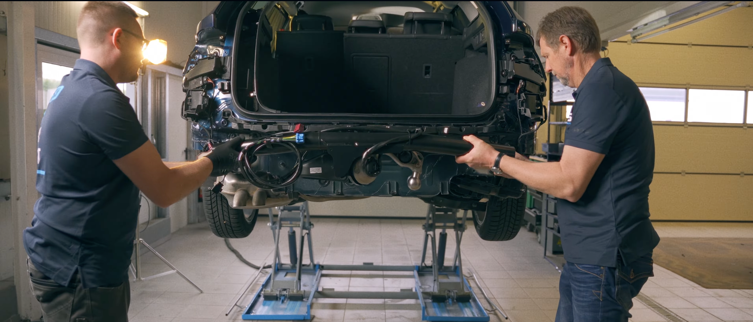 Two technicians installing an ORIS towbar with wiring harness on the rear of a vehicle in a professional workshop.