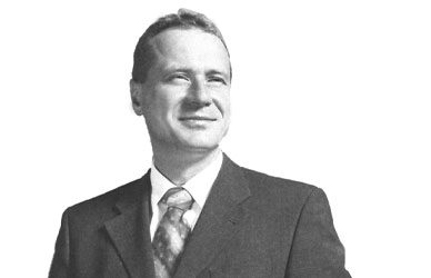 Black-and-white portrait of Jörg Riehle wearing a suit and tie, photographed against a plain light background.