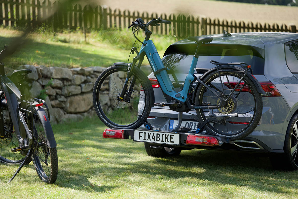 ORIS TRACC FIX4BIKE® bike carrier mounted on a vertically detachable ORIS AK 4 towbar on a grey Volkswagen Golf 8 with an e-bike attached, another bicycle beside the vehicle, and a green landscape with stone wall and wooden fence in the background.