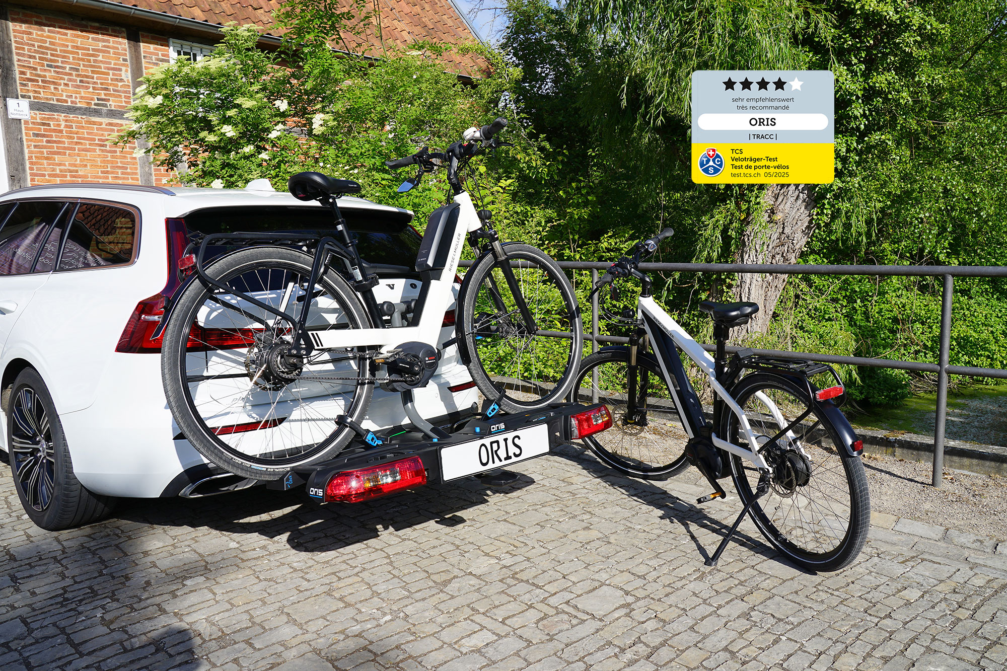 ORIS TRACC FIX4BIKE bicycle carrier mounted on a white Volvo with one e-bike loaded on the carrier, a second e-bike standing beside it and a TCS bicycle carrier test award label in the image, photographed on a cobblestone area with historic buildings