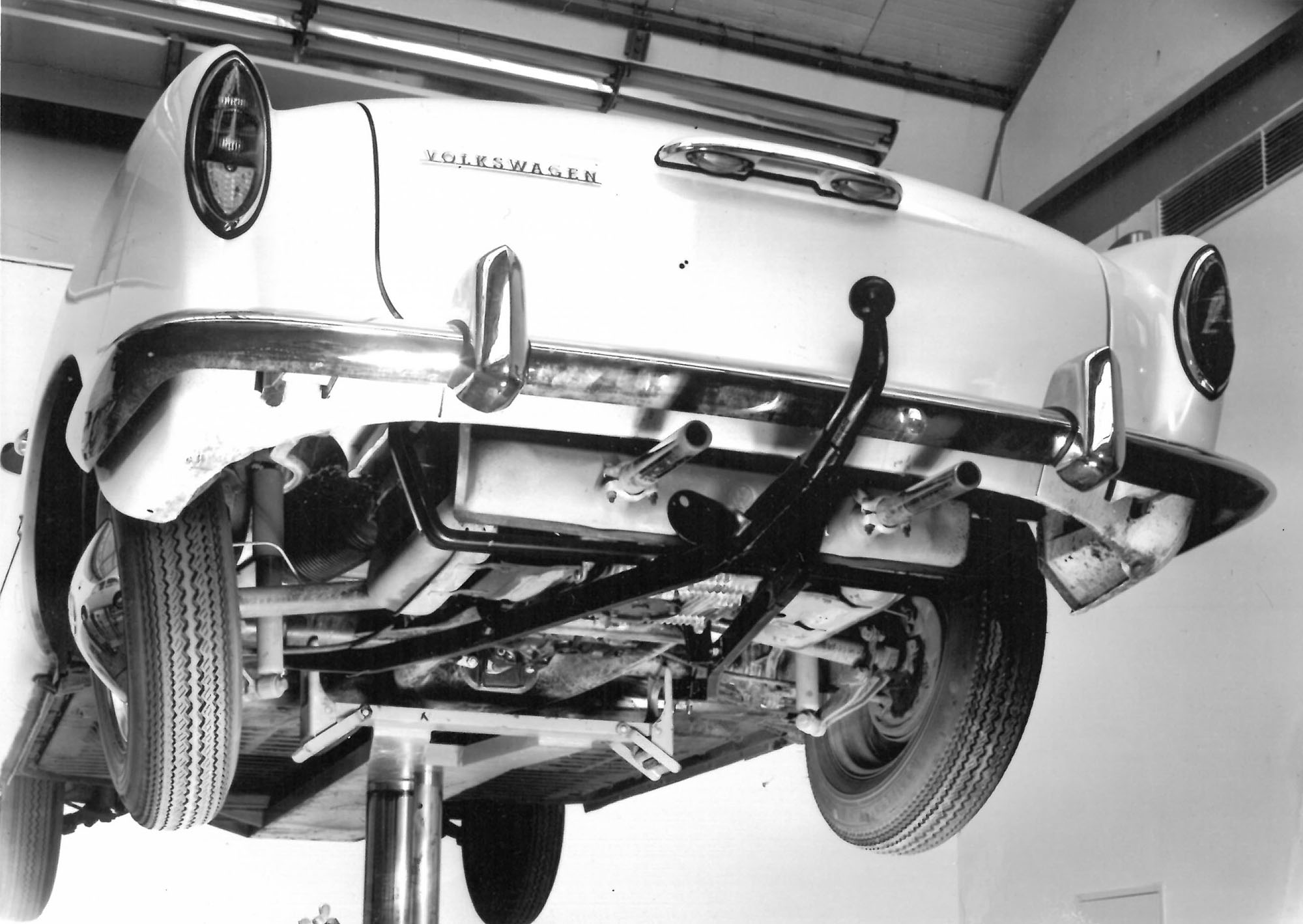 Black-and-white photograph of a classic Volkswagen vehicle on a workshop lift with an early towbar mounted beneath the rear bumper.