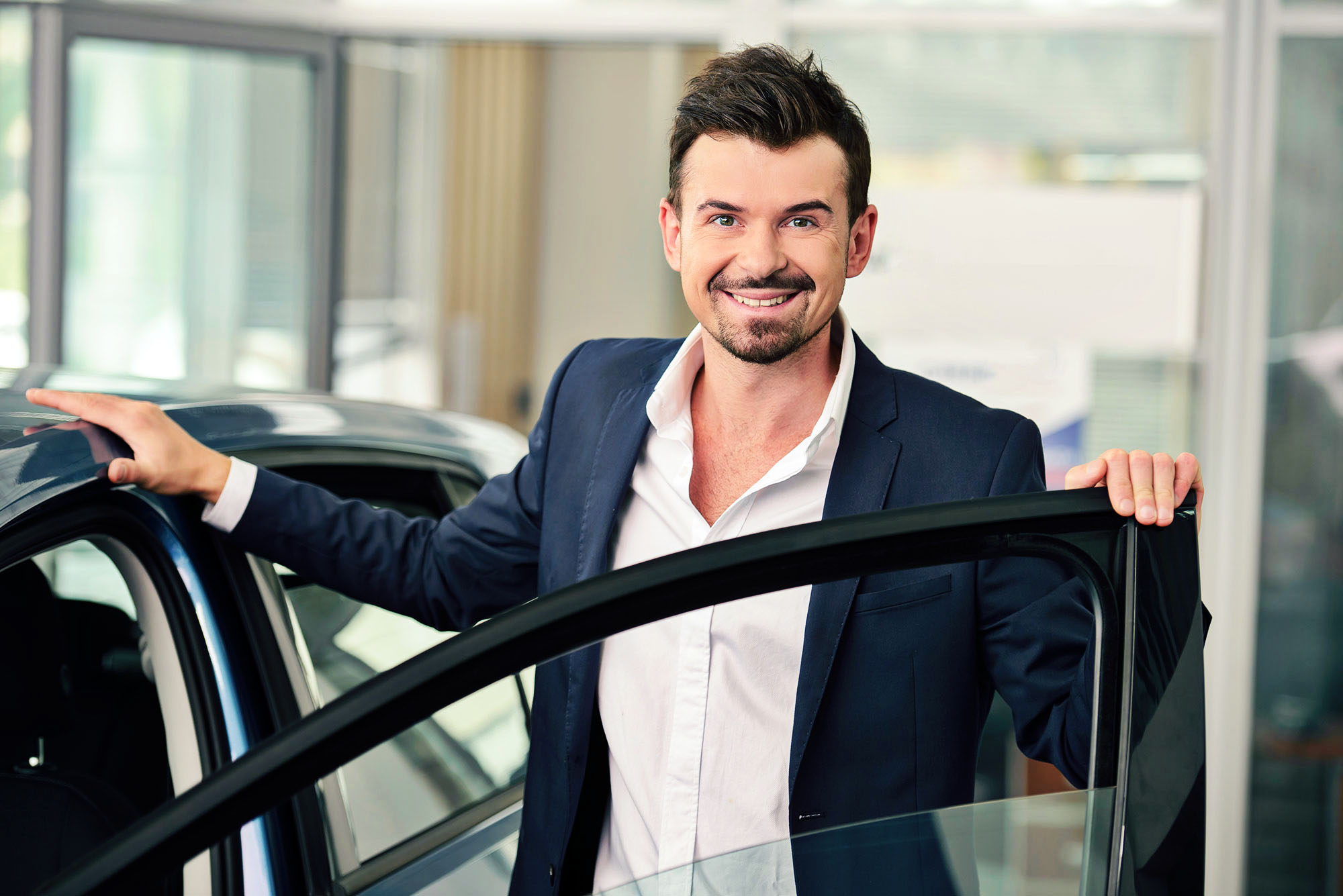 Smiling man standing beside an open car door in a vehicle showroom.