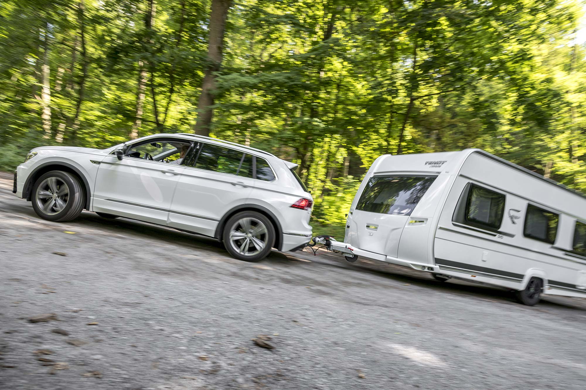 White Volkswagen Tiguan with towbar towing a caravan on a forest road with motion blur and green trees in the background.