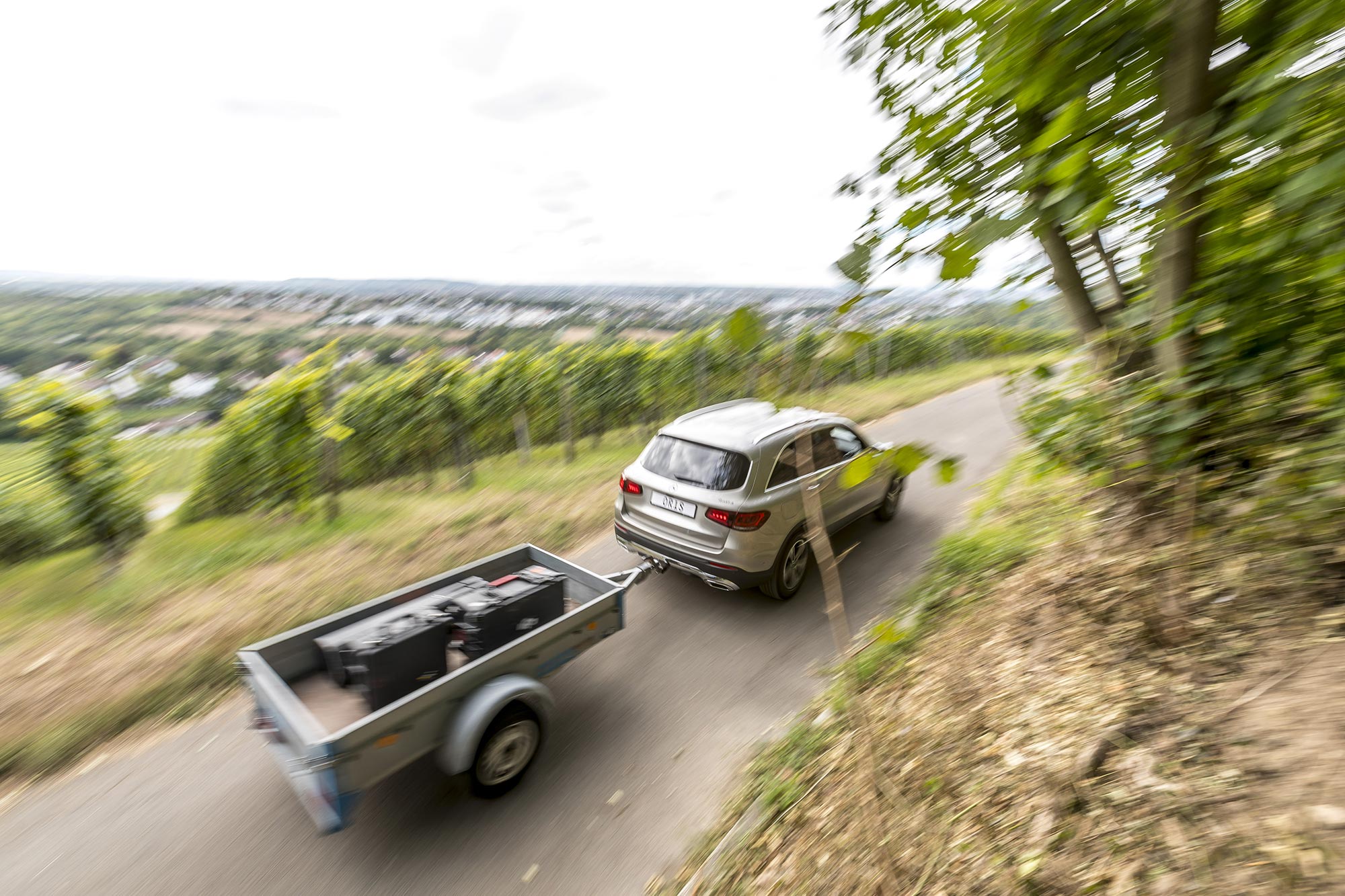 Silver Mercedes-Benz GLC towing a utility trailer on a winding countryside road through vineyards and green hillside landscape.