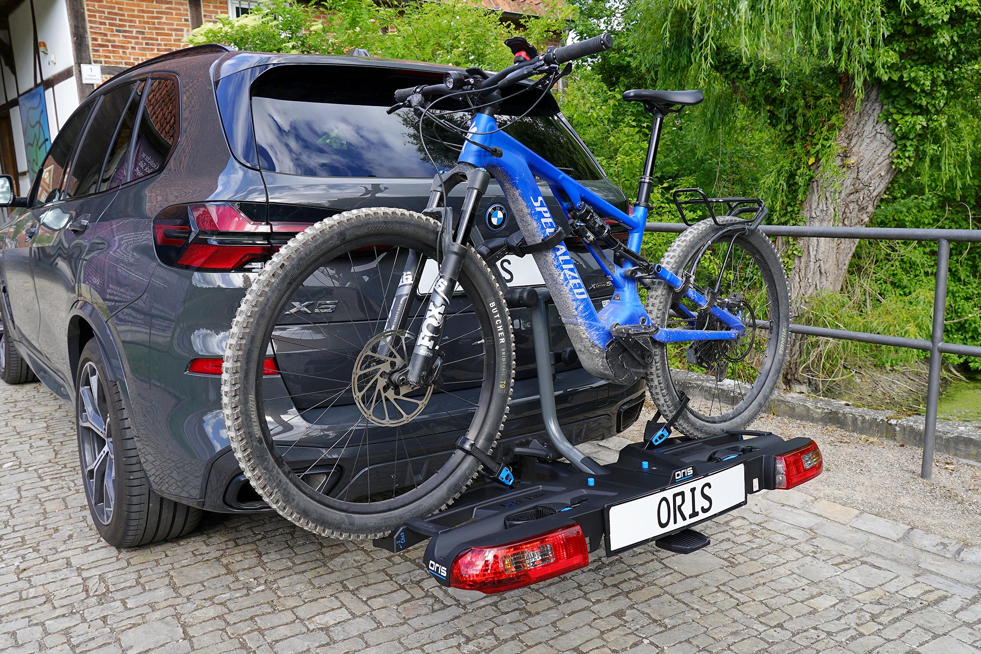 Blue Specialized mountain bike mounted on the ORIS TRACC FIX4BIKE bicycle carrier attached to a dark grey BMW X3, parked on a cobblestone area beside trees and a waterside railing.