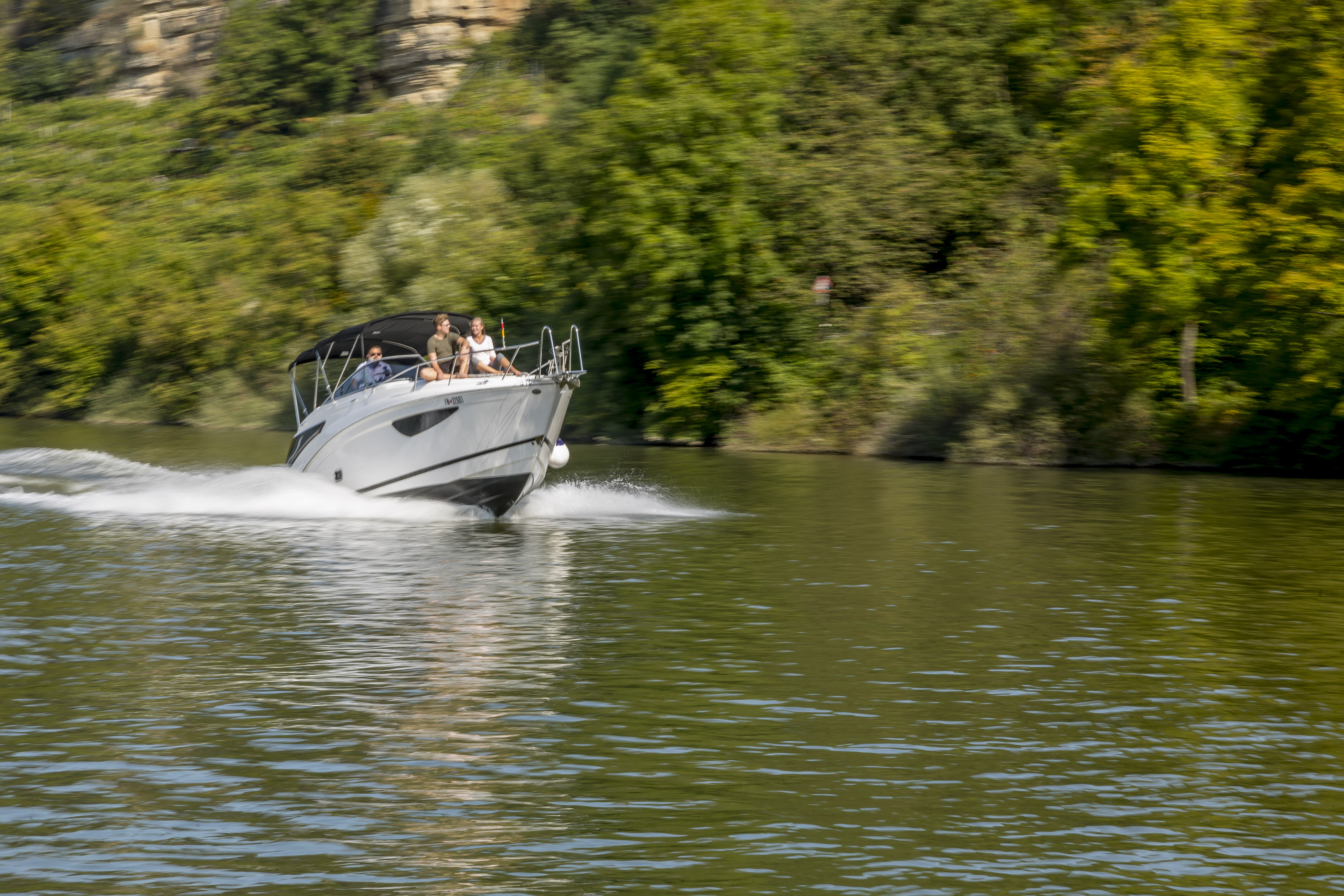 Motorboat cruising on a river with passengers on board, surrounded by green trees and rocky cliffs in a natural landscape.